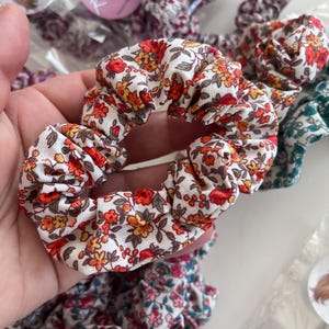 K&ouml;nnte beinhalten: Ein Haargummi aus Stoff mit einem Blumenmuster aus roten, orangefarbenen und gelben Bl&uuml;ten auf wei&szlig;em Grund. Das Haargummi hat ein gerafftes, elastisches Design. Weitere Haargummis mit &auml;hnlichen Blumenmustern sind in der N&auml;he zu sehen.