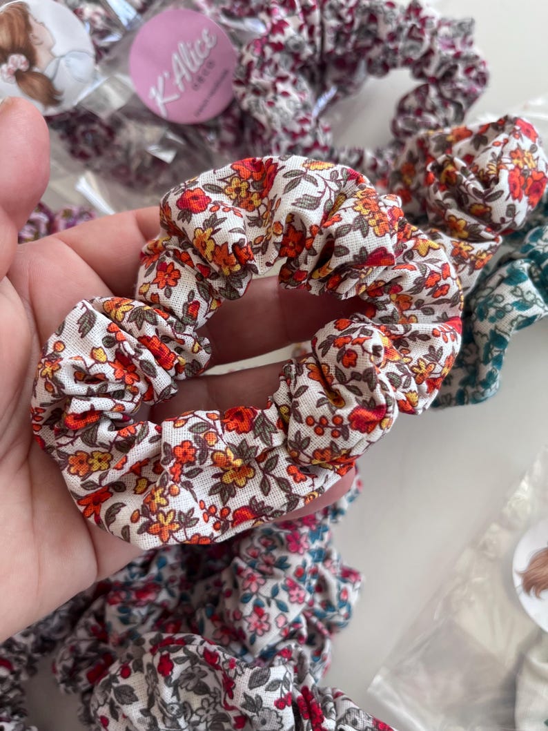 K&ouml;nnte beinhalten: Ein Haargummi aus Stoff mit einem Blumenmuster aus roten, orangefarbenen und gelben Bl&uuml;ten auf wei&szlig;em Grund. Das Haargummi hat ein gerafftes, elastisches Design. Weitere Haargummis mit &auml;hnlichen Blumenmustern sind in der N&auml;he zu sehen.