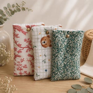 Peut inclure: Trois pochettes rectangulaires en tissu avec fermetures à pression. La première présente un motif toile de Jouy rouge et blanc, la deuxième un quadrillage blanc avec un ours brun et des fleurs, et la troisième un imprimé floral vert et blanc.