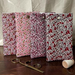 Peut inclure: Quatre étuis en tissu floral rose et rouge, chacun avec une fermeture à bouton-pression. Une paire de ciseaux vintage et une bobine de fil sont posés sur une surface en bois, suggérant un thème de couture ou d'artisanat. Les étuis présentent un motif de petites fleurs.