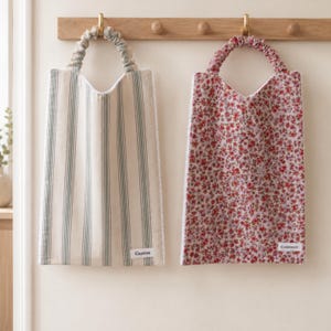 Peut inclure: Deux sacs en tissu rectangulaires avec des anses froncées suspendus à une patère en bois. Le sac de gauche présente des rayures verticales vertes sur fond beige avec une étiquette blanche Gaston. Le sac de droite a un motif floral rouge avec une étiquette Constance.