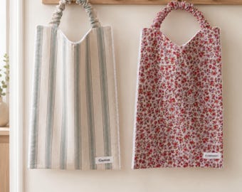 Peut inclure: Deux sacs en tissu rectangulaires avec des anses froncées suspendus à une patère en bois. Le sac de gauche présente des rayures verticales vertes sur fond beige avec une étiquette blanche Gaston. Le sac de droite a un motif floral rouge avec une étiquette Constance.