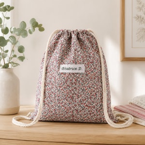 Puede incluir: Una mochila de cordón con un denso estampado floral en tonos rojos, verdes y blancos. La bolsa tiene correas de cuerda gruesas de color crema y una etiqueta rectangular blanca centrada en el frente con el texto Béatrice D. impreso en negro.