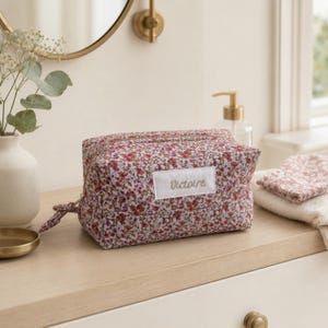 Puede incluir: Un neceser rectangular con un estampado floral rosa y rojo sobre un fondo claro. La bolsa tiene una etiqueta de tela rectangular blanca en la parte delantera con el nombre Victoire bordado en letra cursiva. Incluye un cierre de cremallera.