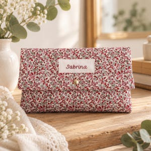 Op de afbeelding: Een rechthoekig stoffen tasje met een dicht bloemenpatroon in rood-, roze- en grijstinten. Een wit rechthoekig label is in het midden op de voorflap geplaatst met de naam Sabrina in het rood geborduurd. Een goudkleurige drukknoop sluit de flap.