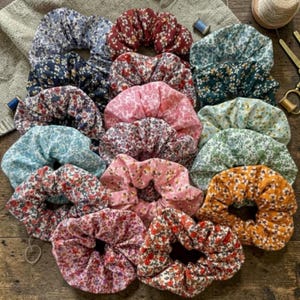 May include: A selection of floral scrunchies in shades of blue, red, pink, and orange. The fabric scrunchies are displayed on a wooden surface. These hair accessories feature a variety of floral patterns and are designed for tying up hair.