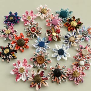 Può includere: Una collezione di fiori in tessuto, ognuno caratterizzato da un bottone centrale. I fiori sono realizzati con vari tessuti a fantasia e a tinta unita, inclusi motivi floreali, geometrici e astratti nelle tonalità del rosso, blu, verde e rosa.