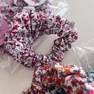 K&ouml;nnte beinhalten: Zwei Haargummis aus Stoff mit Blumenmustern. Das obere Haargummi hat eine wei&szlig;e Basis mit kleinen roten, rosa und grauen Blumenprints. Das untere Haargummi zeigt eine wei&szlig;e Basis mit einem dichten Muster aus kleinen orangen, roten und blauen Blumen.