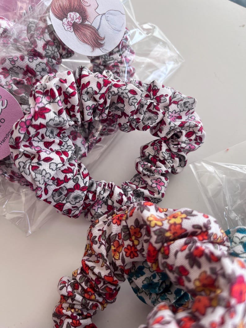 K&ouml;nnte beinhalten: Zwei Haargummis aus Stoff mit Blumenmustern. Das obere Haargummi hat eine wei&szlig;e Basis mit kleinen roten, rosa und grauen Blumenprints. Das untere Haargummi zeigt eine wei&szlig;e Basis mit einem dichten Muster aus kleinen orangen, roten und blauen Blumen.