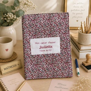 Könnte beinhalten: Ein rechteckiger Notizbuchumschlag mit einem dichten Blumenmuster in Burgunderrot, Rosa und Grau. Ein weißes Stoffetikett ist mittig aufgenäht und trägt den gestickten Text: Mon cahier d'appel, Juliette, Classe de CE1.