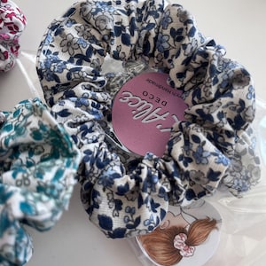 K&ouml;nnte beinhalten: Ein Haargummi aus Stoff mit einem blau-grauen Blumenmuster auf wei&szlig;em Grund. Das Haargummi ist in einer durchsichtigen Plastikt&uuml;te verpackt, die ein rundes rosa Etikett mit der Aufschrift L'Alice DECO und eine kreisf&ouml;rmige Kopfabbildung enth&auml;lt.