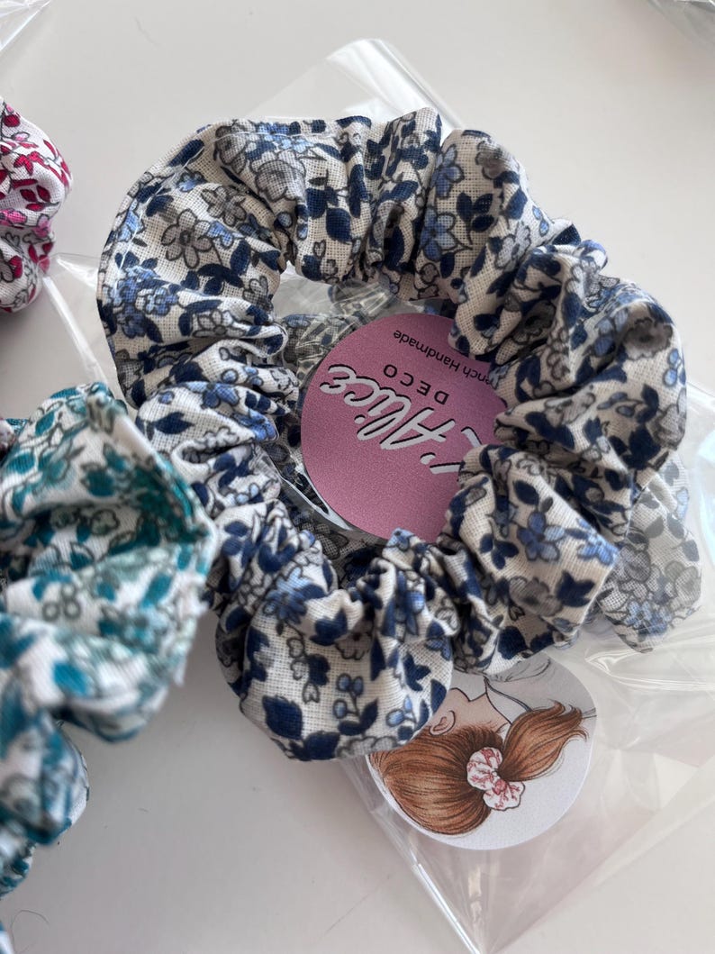 K&ouml;nnte beinhalten: Ein Haargummi aus Stoff mit einem blau-grauen Blumenmuster auf wei&szlig;em Grund. Das Haargummi ist in einer durchsichtigen Plastikt&uuml;te verpackt, die ein rundes rosa Etikett mit der Aufschrift L'Alice DECO und eine kreisf&ouml;rmige Kopfabbildung enth&auml;lt.