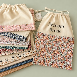 Puede incluir: Una colección de bolsas con cordón hechas de tela color hueso con secciones inferiores de varios patrones que incluyen flores, estampados de animales y cuadros. Una bolsa muestra claramente el texto negro Prénom Brodé en la parte superior.