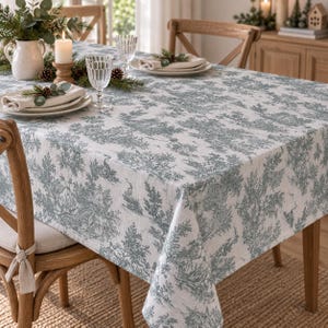 Puede incluir: Un mantel rectangular con base blanca y un patrón toile de jouy azul verdoso que representa árboles, follaje y aves. La tela cubre una mesa de madera dispuesta con servilletas blancas, cristalería transparente y una jarra de cerámica blanca.
