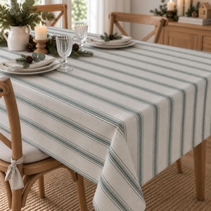 May include: A rectangular tablecloth featuring a pattern of vertical stripes in muted sage green and off-white. The fabric drapes over the edges of a wooden table, which is set with white ceramic plates, clear glass tumblers, and a white pitcher.