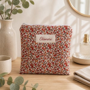 Puede incluir: Un estuche de tela cuadrado con un denso estampado floral de pequeñas flores rojas y naranjas con hojas verdes sobre un fondo crema. Una etiqueta rectangular de color crema está centrada con el nombre Éléonore impreso en rojo.