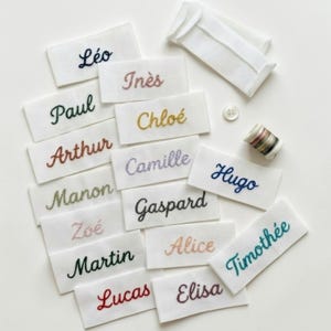 Peut inclure: Étiquettes de noms en tissu blanc brodées avec différents noms en différentes couleurs. Les noms incluent Léo, Paul, Arthur, Manon, Zoé, Martin, Lucas, Inès, Chloé, Camille, Gaspard, Alice, Elisa, Hugo et Timothée. Comprend également un bouton, du fil et une petite pochette en tissu blanc.