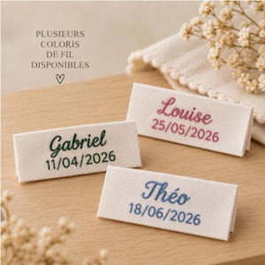 Può includere: Tre etichette rettangolari in tessuto color crema con nomi e date ricamati. Un'etichetta riporta Gabriel 11/04/2026 in verde scuro, Louise 25/05/2026 in rosa e Théo 18/06/2026 in blu. Il testo recita PLUSIEURS COLORIS DE FIL DISPONIBLES.