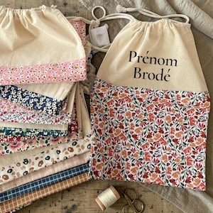 Peut inclure: Sacs à cordon beiges avec des fonds à motifs floraux. Un sac porte le texte "Prénom Brodé". Une variété de motifs floraux sont visibles, notamment des motifs floraux roses, bleus et rouges. Une bobine de fil et des ciseaux sont au premier plan.