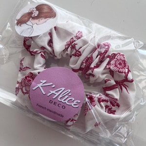 K&ouml;nnte beinhalten: Ein wei&szlig;es Stoff-Haargummi mit einem dunkelrosa Blumenmuster. Das Haargummi ist in einer transparenten Kunststoffh&uuml;lle verpackt, die mit einem runden Etikett mit einer Personenzeichnung und einem lila Aufkleber mit dem Text K'Alice DECO versehen ist.