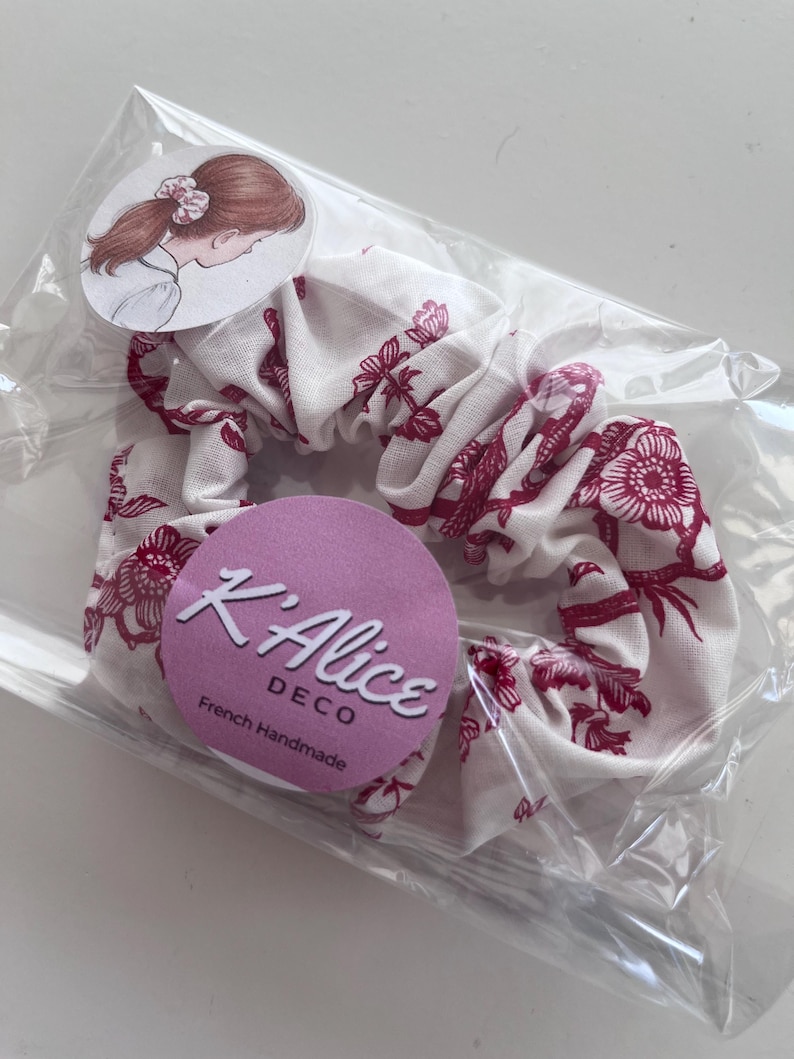 K&ouml;nnte beinhalten: Ein wei&szlig;es Stoff-Haargummi mit einem dunkelrosa Blumenmuster. Das Haargummi ist in einer transparenten Kunststoffh&uuml;lle verpackt, die mit einem runden Etikett mit einer Personenzeichnung und einem lila Aufkleber mit dem Text K'Alice DECO versehen ist.