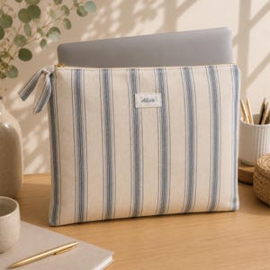 Peut inclure: Une housse d'ordinateur portable rectangulaire en tissu, dotée d'une base couleur crème avec des rayures verticales bleues. Elle possède une tirette de fermeture éclair assortie et une étiquette blanche avec le nom Alice écrit en noir.