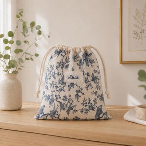 Può includere: Una borsa in tessuto color crema con chiusura a coulisse e un motivo botanico e di uccelli blu. Un'etichetta bianca rettangolare è cucita al centro della borsa con il nome Alice stampato in un carattere corsivo blu scuro.