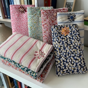May include: A selection of fabric book covers and cushions in floral and striped designs. The covers have decorative flower embellishments. Colours include pink, blue, red, and white. The cushions are stacked on a white shelf.