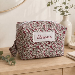 Peut inclure: Une trousse de toilette rectangulaire en tissu avec un motif floral dense dans des tons de rouge, vert et blanc. Une étiquette rectangulaire blanche est centrée sur le devant avec le prénom "Éléonore" brodé en rouge. Fermeture éclair blanche.