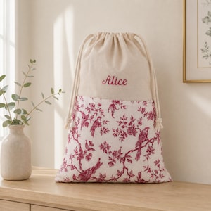 Op de afbeelding: Een tas met trekkoord van gebroken wit textiel met een rood-wit toile-patroon van vogels en bloemtakken op de onderste helft. Het bovenste gedeelte is van effen stof met de naam Alice in rode cursieve letters geborduurd.