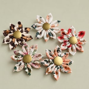 Può includere: Cinque decorazioni a forma di fiore in tessuto con centri in metallo color ottone. Ogni fiore presenta motivi distinti tra cui stampa leopardata, bianco con foglie verdi e disegni floreali rossi, rosa e arancioni con petali piegati.
