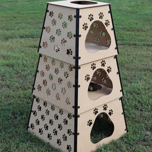 Puede incluir: Una torre de gato de madera de tres niveles con recortes de huellas de patas. Cada nivel tiene una abertura circular para que los gatos puedan entrar. La torre está hecha de madera marrón y tiene detalles negros.