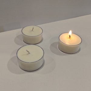 May include: Three white tea light candles, one of which is lit, on a white surface.