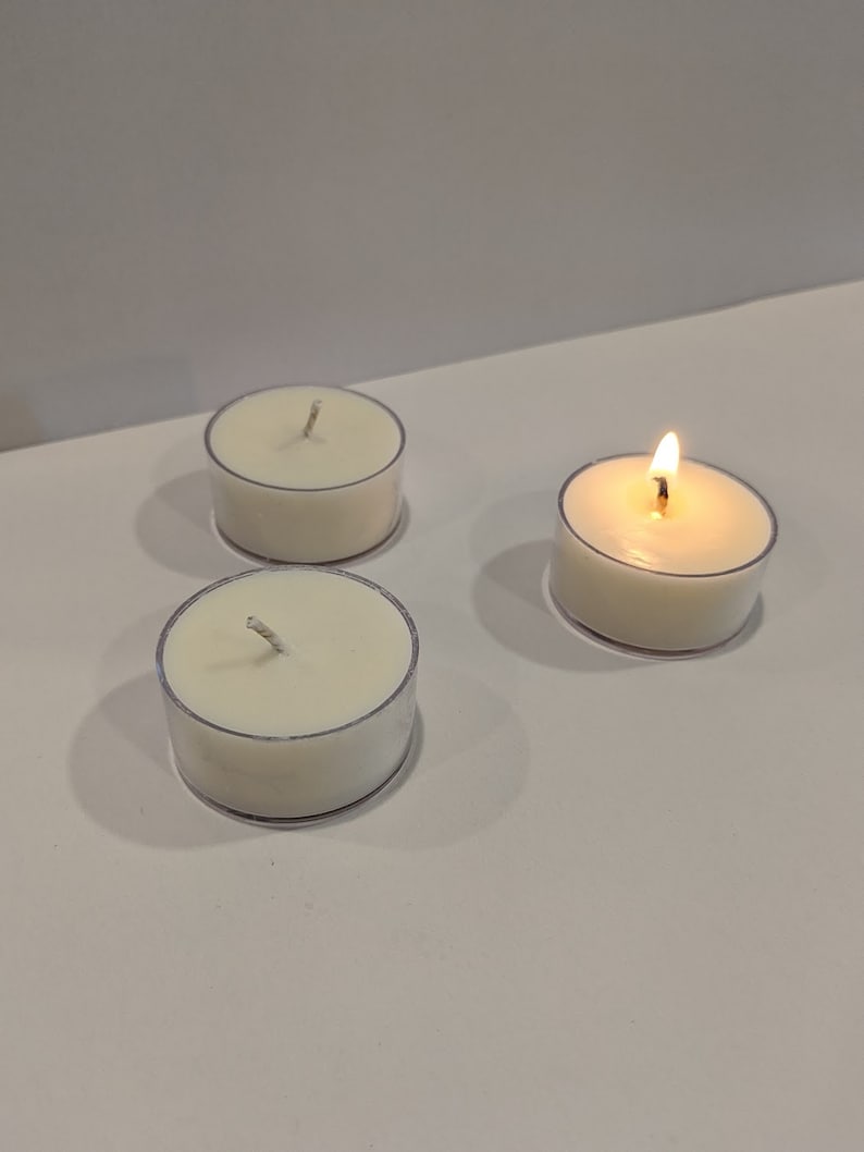 May include: Three white tea light candles, one of which is lit, on a white surface.