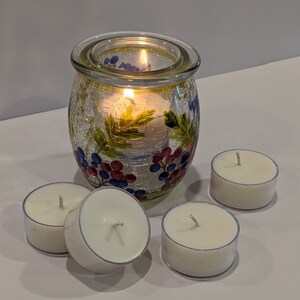 May include: A set of four white tea light candles and a glass candle holder with a floral design. The candle holder has a textured surface and is decorated with blue, red, and green painted grapes and leaves.