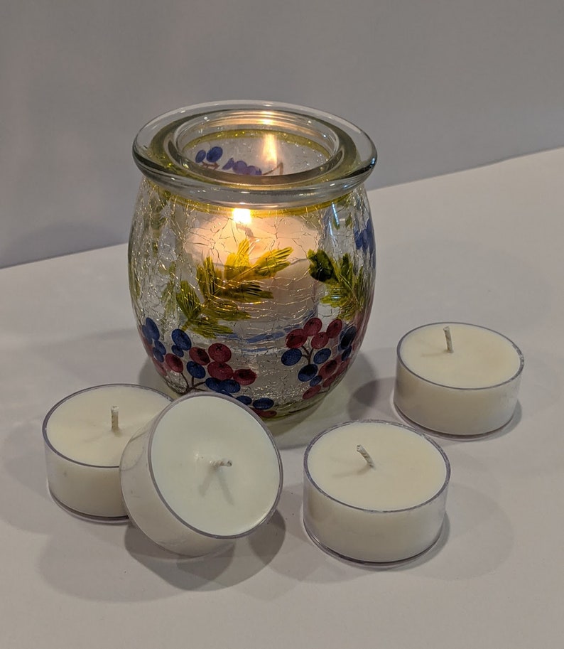 May include: A set of four white tea light candles and a glass candle holder with a floral design. The candle holder has a textured surface and is decorated with blue, red, and green painted grapes and leaves.