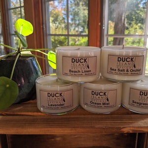 May include: Five glass candle jars with white wax and brown labels. The labels have the text "Duck Island Designs" and the scent names: "Beach Linen", "Sea Salt & Orchid", "Ocean Mist", "Seagrass & Sage", and "Lavender".