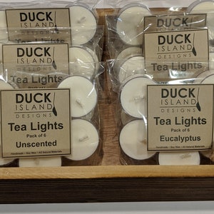 May include: A wooden box filled with six packs of unscented and eucalyptus scented tea lights. Each pack contains six tea lights and is labeled "Duck Island Designs Tea Lights".