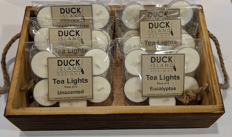May include: A wooden box filled with six packs of unscented and eucalyptus scented tea lights. Each pack contains six tea lights and is labeled "Duck Island Designs Tea Lights".