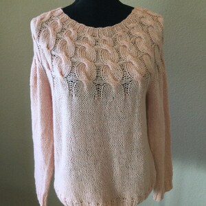 May include: A pale pink knitted sweater with a cable knit design around the neckline. The sweater has long sleeves and a relaxed fit.