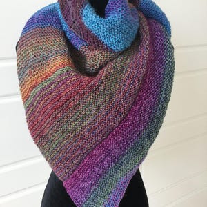 May include: A hand-knitted shawl in a triangular shape, featuring a gradient of colors from orange to purple and blue. The shawl is displayed on a black mannequin, showcasing its textured knit and vibrant hues.