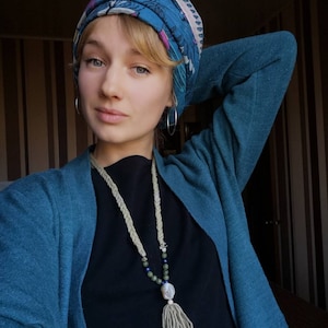 May include: A woman wearing a blue cardigan and a black top with a long, white, beaded necklace with a tassel. She is wearing a patterned headscarf.