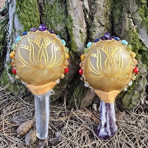 May include: Two decorative wands with a golden lotus design on a beige orb, adorned with colorful gemstones. One wand has a clear crystal handle, the other a purple crystal handle. The background features a tree trunk and pine needles.