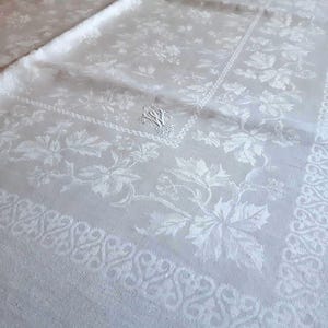 Pode incluir: Toalha de mesa branca com um padrão floral e de folhas. O design inclui uma borda com um padrão de rolagem repetido. Um monograma é visível no centro. O tecido parece ser um material macio e tecido.