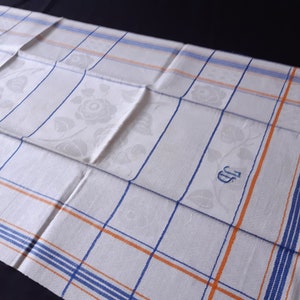 May include: White linen kitchen towel with a blue and orange checked pattern and a floral design. The towel has the initials "JS" embroidered in the centre.