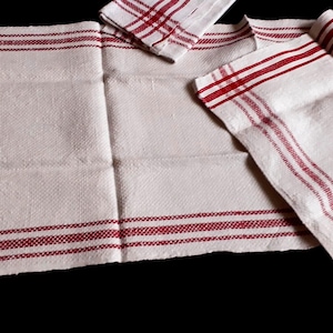 May include: White linen kitchen towels with red stripes. The towels are folded and arranged. The design includes a red border and stripes across the fabric. The set includes a small square napkin. The background is black.