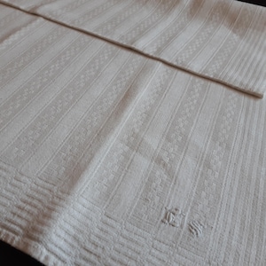 Pode incluir: Uma toalha de mesa de linho branco com um padrão em relevo sutil e uma inicial bordada no canto inferior direito.