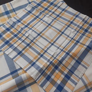 Pode incluir: Tecido com padrão xadrez branco, azul e amarelo. O tecido é dobrado e disposto em uma composição em camadas. O design xadrez apresenta linhas que se cruzam em azul, amarelo e branco, criando um visual clássico e versátil. O tecido parece ser de algodão ou material semelhante.