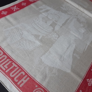 Könnte beinhalten: Eine weiße Leinen-Tischdecke mit rotem Rand und dem in Rot gedruckten Wort "Rolltuch". Die Tischdecke zeigt ein weiß gesticktes Design einer Gruppe von Menschen.