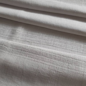 Pode incluir: Tecido de linho branco com uma trama texturizada. O tecido está dobrado e mostra a textura da trama.